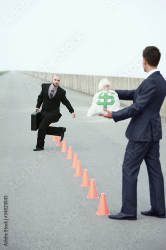 Photo: Businessman running in between traffic cones, another businessman holding a bag of money Businessman running in between traffic cones, another businessman holding a bag of money