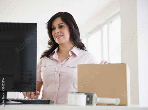 Photo: Smiling Hispanic woman with cardboard box using computer Smiling Hispanic woman with cardboard box using computer