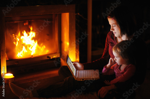 Photo: Young mother and daughter by a fireplace with a laptop Young mother and daughter by a fireplace with a laptop