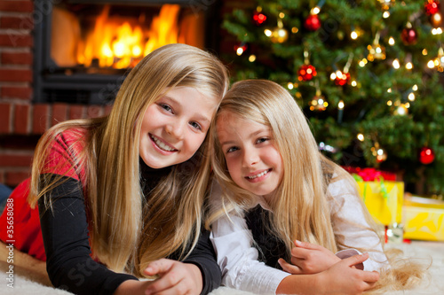 Photo: two girls on christmas eve tree and fire place in background two girls on christmas eve tree and fire place in background
