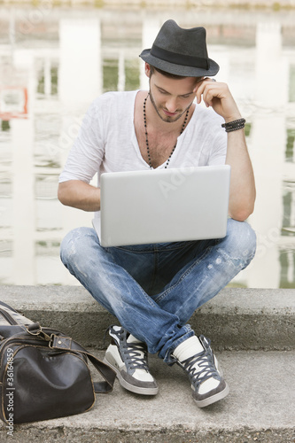Photo: Man sitting on the ledge of a canal and using a laptop, Paris, Ile-de-France, France Man sitting on the ledge of a canal and using a laptop, Paris, Ile-de-France, France