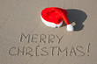 Photo: Merry Christmas written on the sand beach under red Santa hat Merry Christmas written on the sand beach under red Santa hat