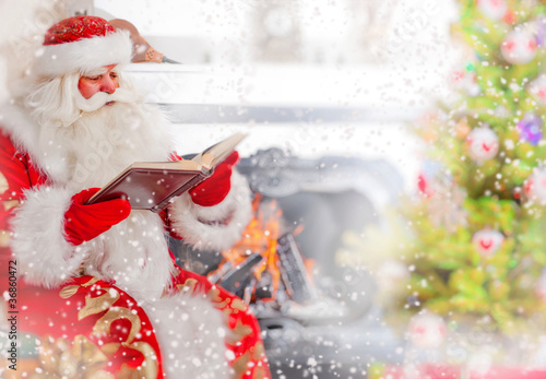 Photo: Santa sitting at the Christmas tree, near fireplace and reading Santa sitting at the Christmas tree, near fireplace and reading