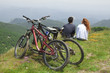 Couple sitting together on hill, looking at view, bicycles parked behind them
