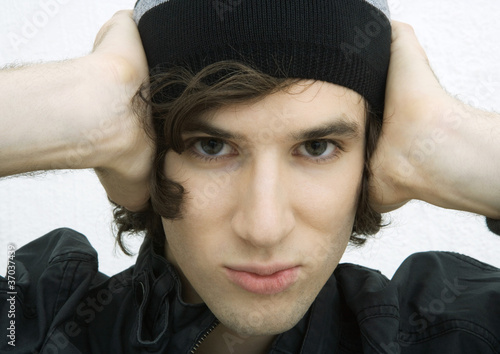 Photo: Young man with hands on side of head, looking at camera, close-up portrait Young man with hands on side of head, looking at camera, close-up portrait