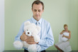 New father holding teddy bear, looking down, wife holding baby in background