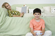 Young boy sitting on floor playing video game, mother relaxing on sofa