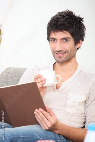 Photo: young man reading a book and drinking coffee on a couch young man reading a book and drinking coffee on a couch