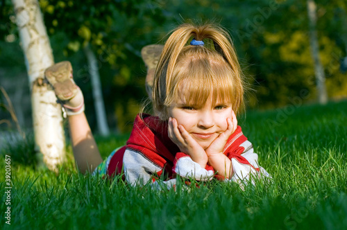 Photo: The small beautiful girl sits on a green lawn The small beautiful girl sits on a green lawn