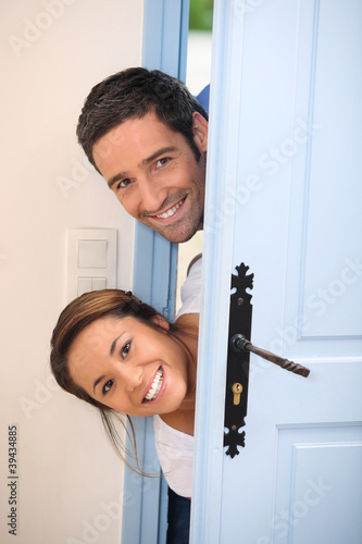 Photo: Young couple poking their heads around the front door Young couple poking their heads around the front door