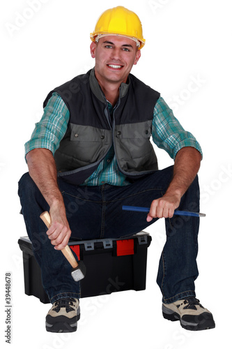 Photo: Smiling tradesman sitting on his toolbox and holding tools Smiling tradesman sitting on his toolbox and holding tools