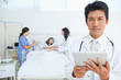 Photo: Serious doctor looking ahead while holding a tablet as his colleagues treat a patient Serious doctor looking ahead while holding a tablet as his colleagues treat a patient