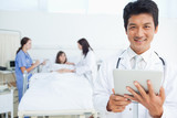 Photo: Doctor looking ahead while holding a tablet as his colleagues treat a patient Doctor looking ahead while holding a tablet as his colleagues treat a patient