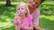 A girl repeatedly blows bubbles with her mother as she sits in the park
