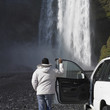 Man in winter jacket leaning on truck and looking at waterfall