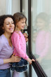 Smiling mother and daughter look out window; woman hugs girl