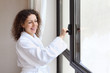 Beautiful smiling woman dressed in white bathrobe opens window