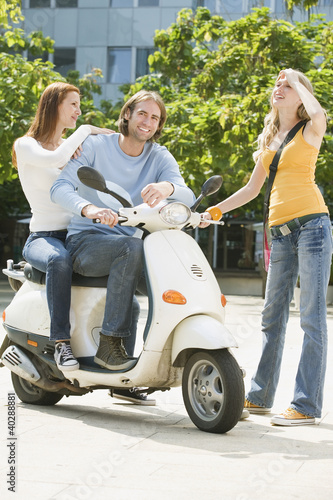 Photo: Young couple sitting on motor scooter while friend smiling Young couple sitting on motor scooter while friend smiling
