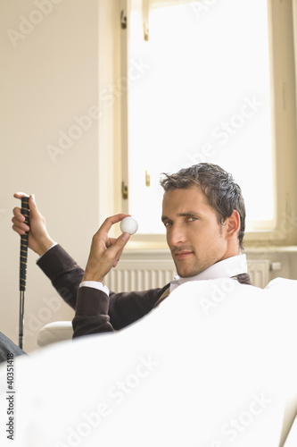 Photo: Mid adult man sitting on sofa, holding golf ball Mid adult man sitting on sofa, holding golf ball