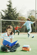 Girl reading while her friends are playing soccer.