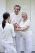 Woman giving senior woman a bouquet of flowers, senior man smiling while watching