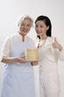 Photo: Senior woman with apron holding bamboo steamers, woman posing beside her showing thumbs up Senior woman with apron holding bamboo steamers, woman posing beside her showing thumbs up