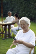 Photo: Senior woman text messaging on the phone, woman having tea with senior man in the background Senior woman text messaging on the phone, woman having tea with senior man in the background