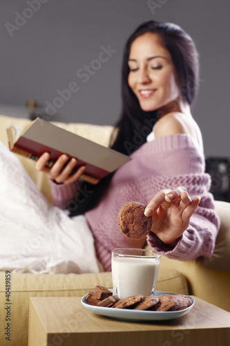 Photo: Woman dipping cookie into a glass of milk while reading book on the couch Woman dipping cookie into a glass of milk while reading book on the couch