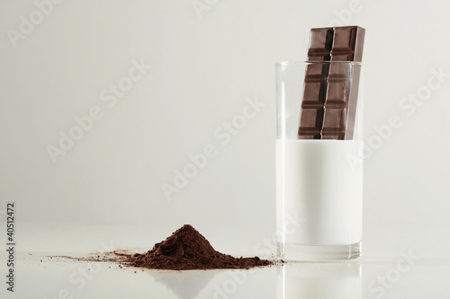 Photo: Chocolate bar dipping into a glass of milk, cocoa powder on the table Chocolate bar dipping into a glass of milk, cocoa powder on the table