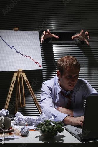 Foto: Businessman frowning while using laptop, hands coming out from window blinds Businessman frowning while using laptop, hands coming out from window blinds