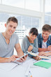 Two students use a book while the student using a tablet looks a