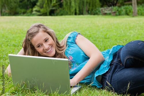 Foto: Young smiling girl lying on the side in a park while typing on h Young smiling girl lying on the side in a park while typing on h
