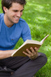 Young smiling man reading a book while siting on the grass