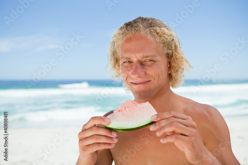 Photo: Smiling blonde man holding a piece of a watermelon while standin Smiling blonde man holding a piece of a watermelon while standin