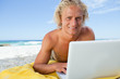 Smiling blonde man lying on the beach while using his laptop