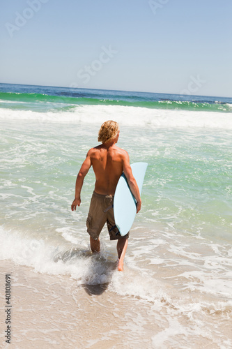 Foto: Young blonde man holding his surfboard while walking in the wate Young blonde man holding his surfboard while walking in the wate