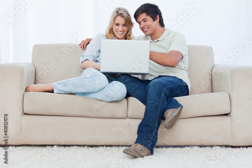 Photo: A smiling couple are sitting on a couch together looking at a la A smiling couple are sitting on a couch together looking at a la