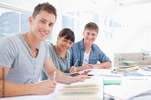 Foto: Three smiling students doing homework as they look into the came Three smiling students doing homework as they look into the came