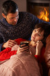 Husband Nursing Sick Wife With Cold Resting On Sofa By Cosy Log