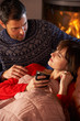 Husband Nursing Sick Wife With Cold Resting On Sofa By Cosy Log