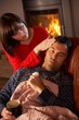 Wife Nursing Sick Husband With Cold Resting On Sofa By Cosy Log