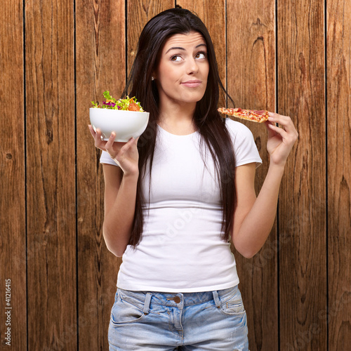 Photo: portrait of young woman choosing pizza or salad against a wooden portrait of young woman choosing pizza or salad against a wooden