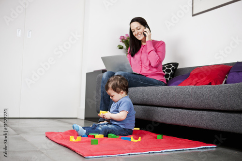 Photo: A mother using a laptop and phone, baby playing A mother using a laptop and phone, baby playing