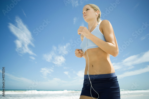 Photo: Young woman jogging on beach, listening to MP3 player, low angle view Young woman jogging on beach, listening to MP3 player, low angle view