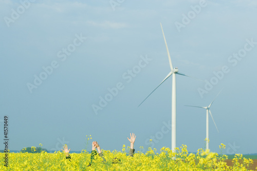 Photo: Two friends hiding in field of colza with arms raised, holding hands, wind turbines in background Two friends hiding in field of colza with arms raised, holding hands, wind turbines in background