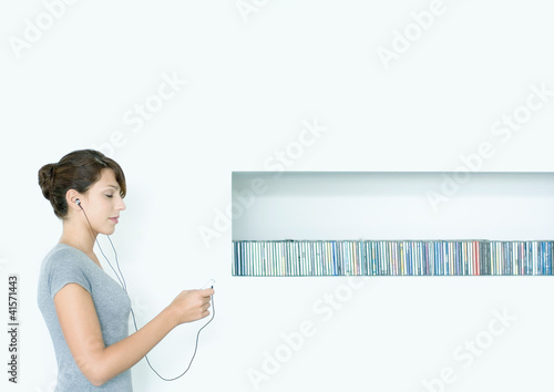 Photo: Young woman standing, listening to MP3 player, next to row of CDs, eyes closed Young woman standing, listening to MP3 player, next to row of CDs, eyes closed