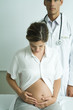 Pregnant woman sitting on examination table, looking down at stomach, doctor standing behind her