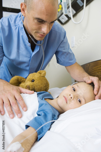 Photo: Girl lying in hospital bed, male nurse's hand on girl's forehead Girl lying in hospital bed, male nurse's hand on girl's forehead