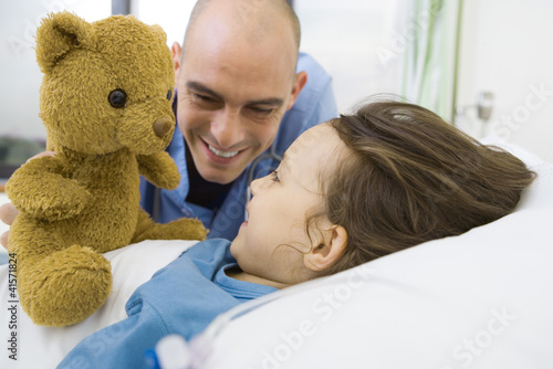 Photo: Girl lying in hospital bed, nurse holding up teddy bear Girl lying in hospital bed, nurse holding up teddy bear