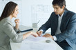 Photo: Female real estate agent pointing to document, man signing, blueprints in foreground and background Female real estate agent pointing to document, man signing, blueprints in foreground and background